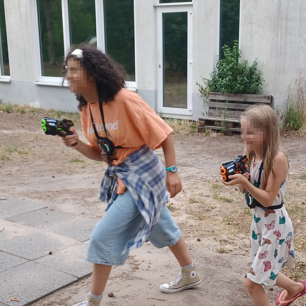 Zwei spielende Kinder, eines älteren und eines jüngeren, laufen draußen neben einem Gebäude und halten bunte Spielzeugblaster.