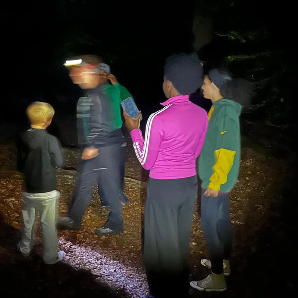 Fünf Kinder stehen nachts auf einem Waldweg, beleuchtet von einer Kopflampe. Eine in pinker Jacke hält ein Smartphone, eine andere trägt eine grün-gelbe Kapuzenjacke, eine Person mit Stirnlampe geht gerade vorbei.