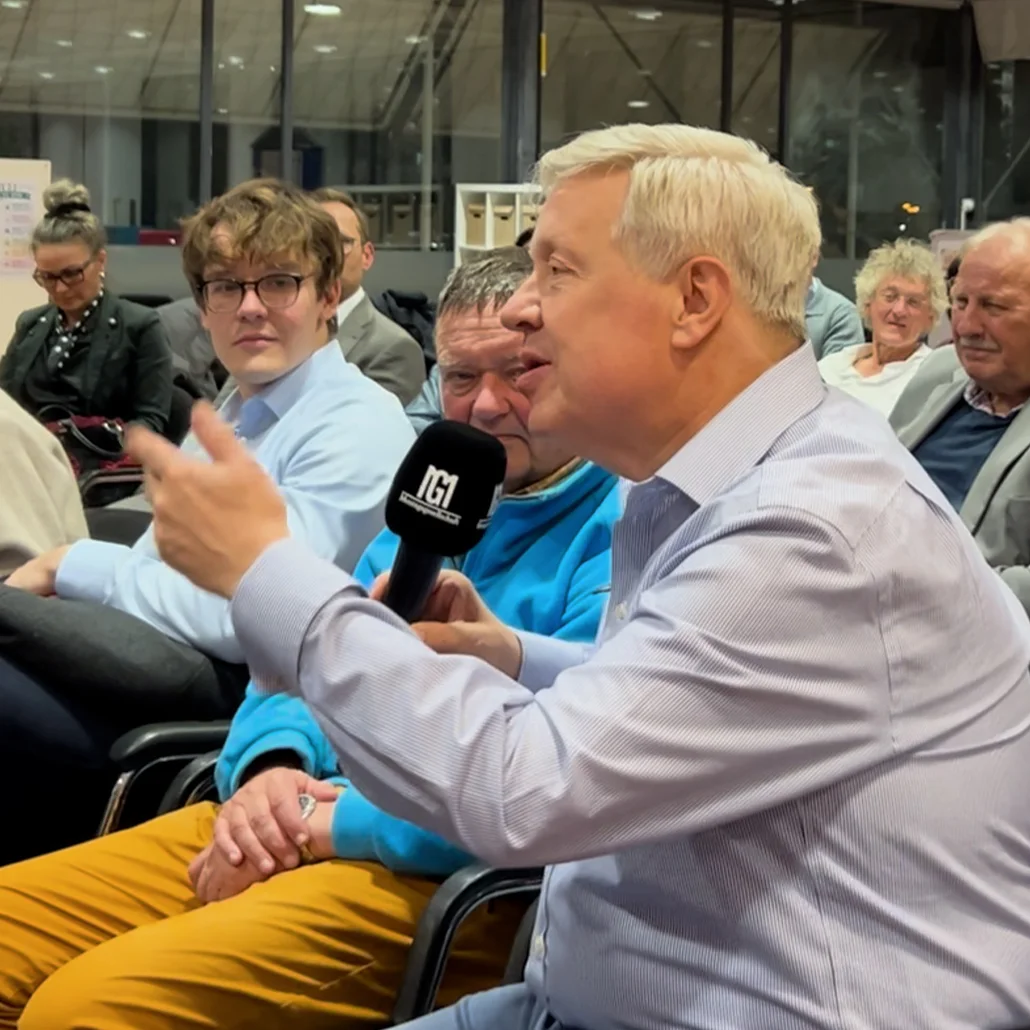 Älterer Mann hält ein Mikrofon und spricht während einer öffentlichen Diskussionsrunde; mehrere Zuhörer sitzen um ihn herum und hören aufmerksam zu.
