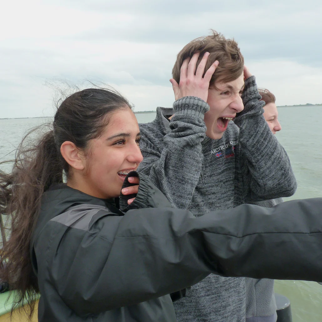 Drei Jugendliche auf einem Boot: eine junge Frau zeigt lachend nach vorn, ein Junge hält sich aufgeregt den Kopf und ruft, dahinter ein weiteres Gesicht, offenes Meer unter bedecktem Himmel.