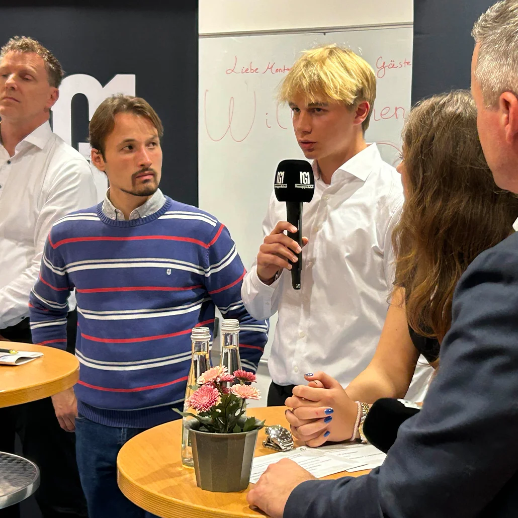 Junger Mann hält ein Mikrofon und spricht an einem Stehtisch, mehrere Menschen hören aufmerksam zu; auf dem Tisch stehen Blumen, Wasserflaschen und Unterlagen.