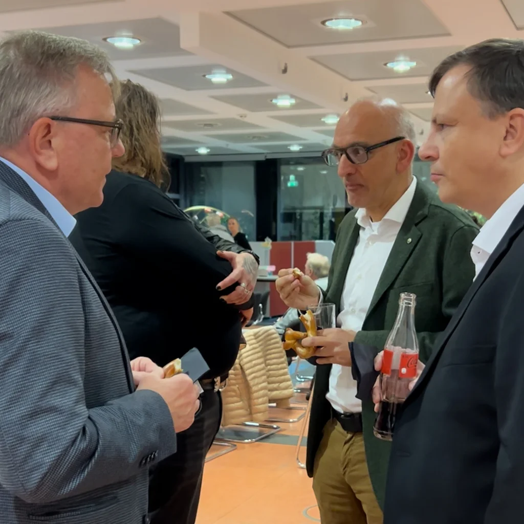 Drei Männer in Jacken stehen bei einer Abendveranstaltung in einem Innenraum im Gespräch; sie essen Häppchen und trinken, einer hält eine Cola‑Flasche.