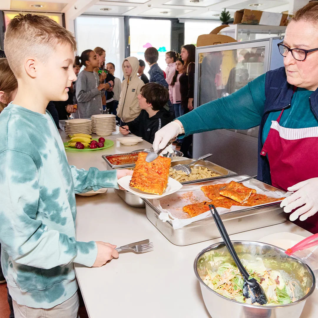 Schulkantine: Eine Mitarbeiterin mit Schürze und Handschuhen serviert einem Jungen ein Stück Lasagne; weitere Kinder stehen in der Essensschlange.