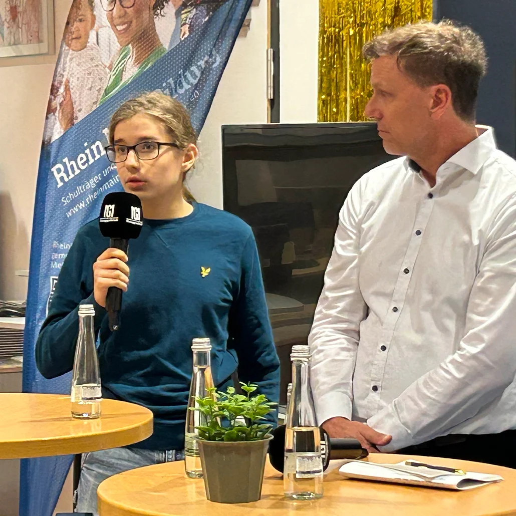 Zwei Personen bei einer Diskussionsrunde: eine junge Person mit Brille hält ein Mikrofon und spricht, ein Mann in weißem Hemd hört zu. Auf Tischen Wasserflaschen und eine kleine Topfpflanze; Banner im Hintergrund.