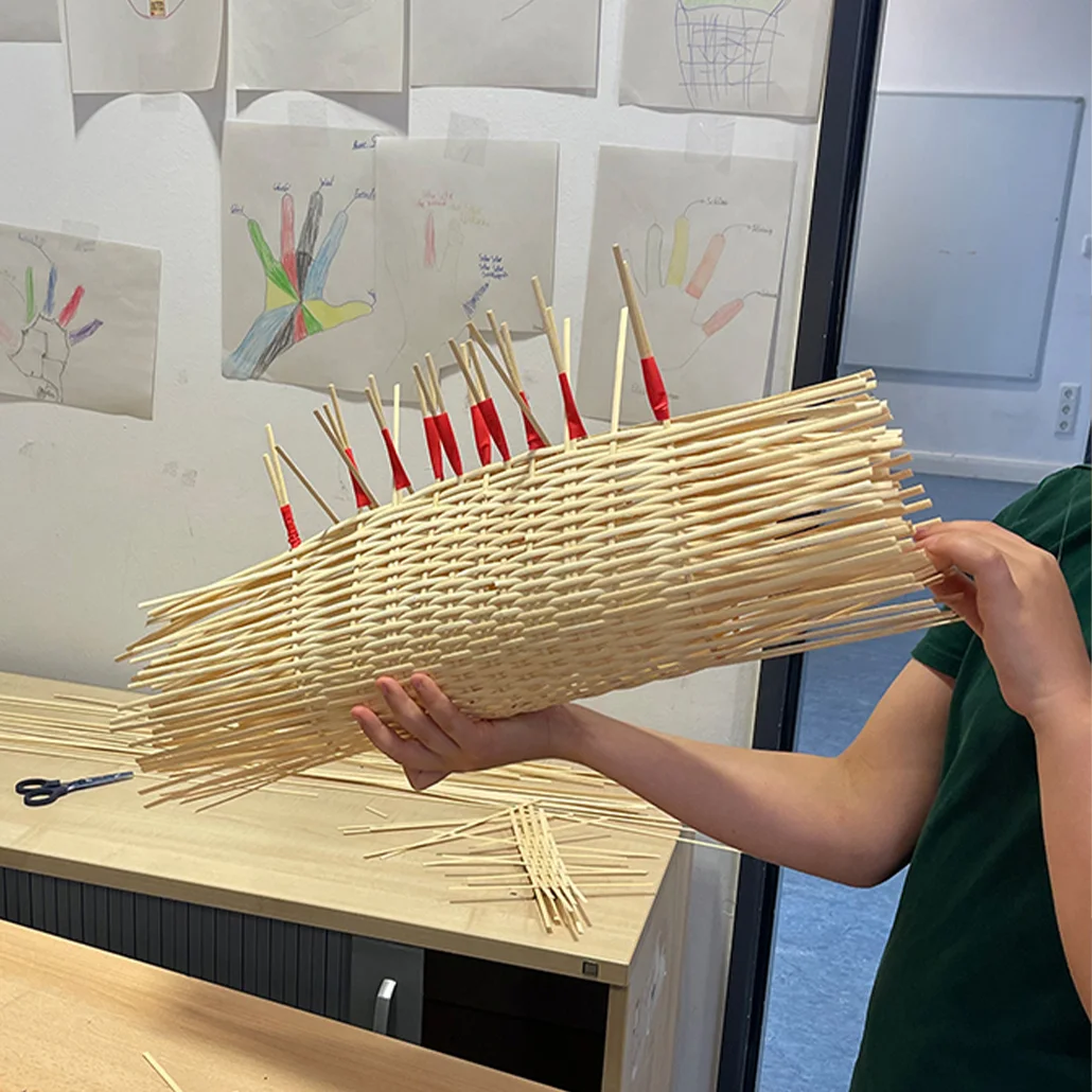 Handgefertigtes Geflecht aus vielen Holzspießen mit roten Spitzen, zu einem länglichen Korb geformt und von zwei Händen gehalten, offenbar ein Bastelprojekt im Klassenraum.