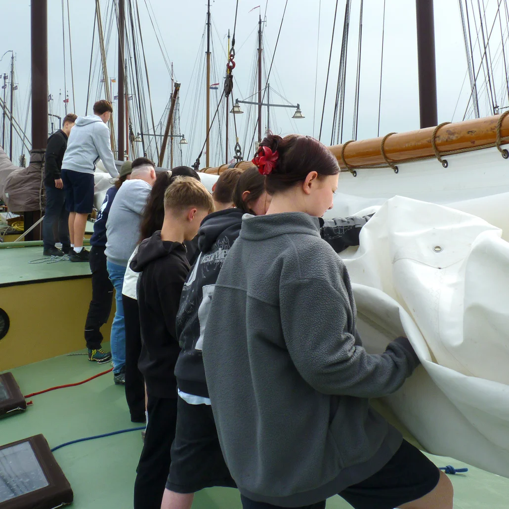 Mehrere Jugendliche stehen an Deck eines Segelschiffs und ziehen gemeinsam ein großes Segeltuch/eine Segelplane, im Hintergrund zahlreiche Masten.