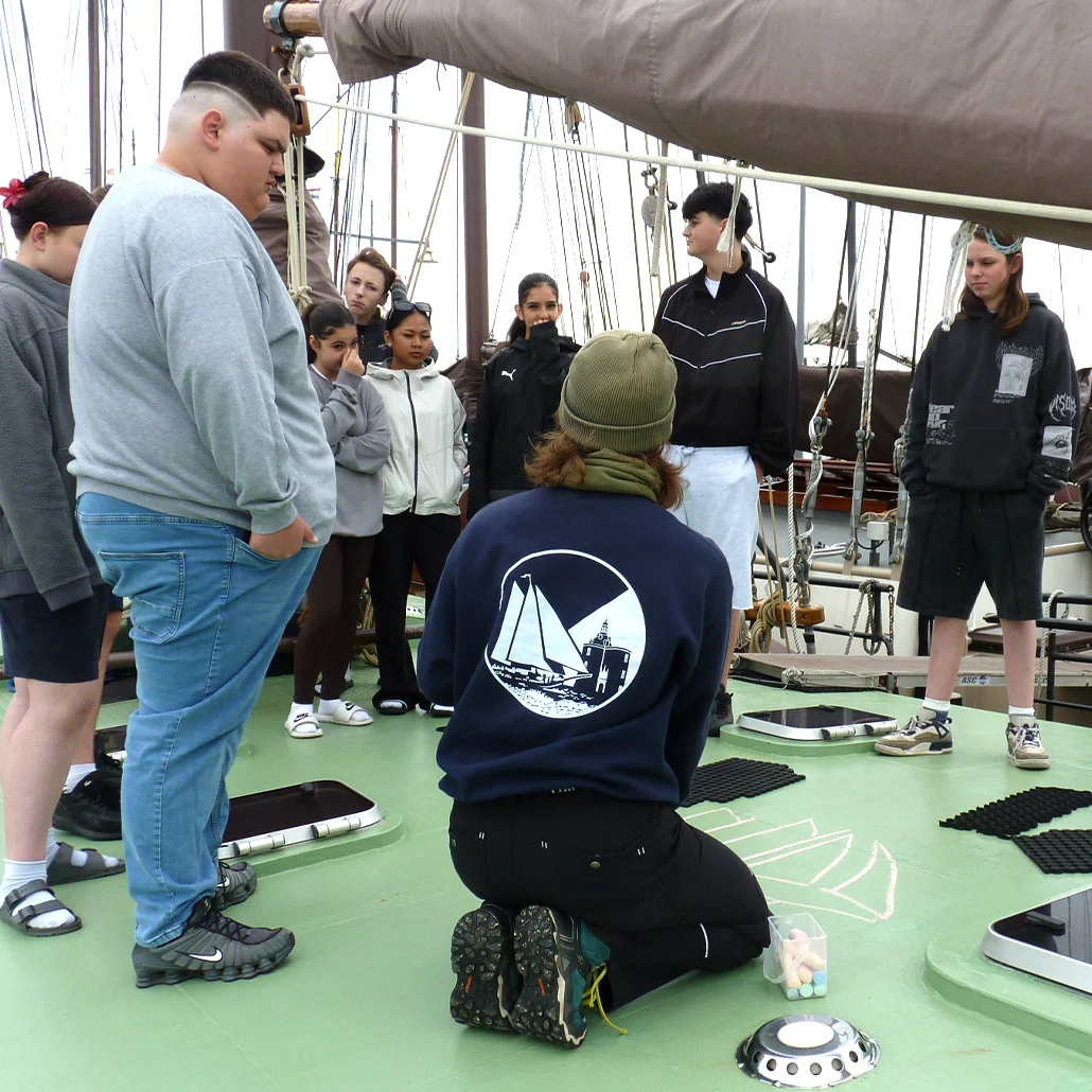 Gruppe Jugendlicher steht auf dem Deck eines Segelschiffs, eine Person kniet vor ihnen und instruiert; Takelage und Segel im Hintergrund.