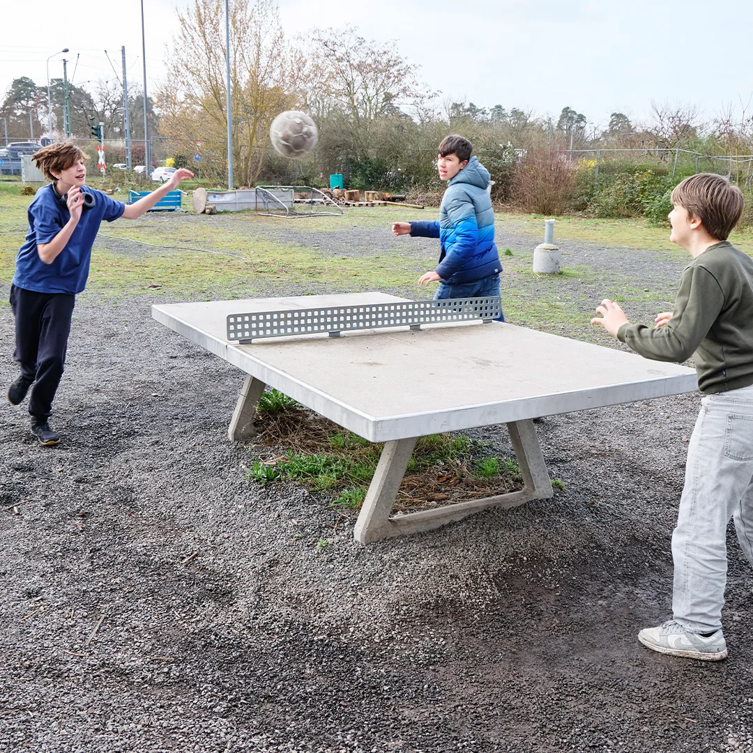 Drei Jungen spielen draußen an einem Beton-Tischtennistisch und schlagen einen Fußball über das Netz.