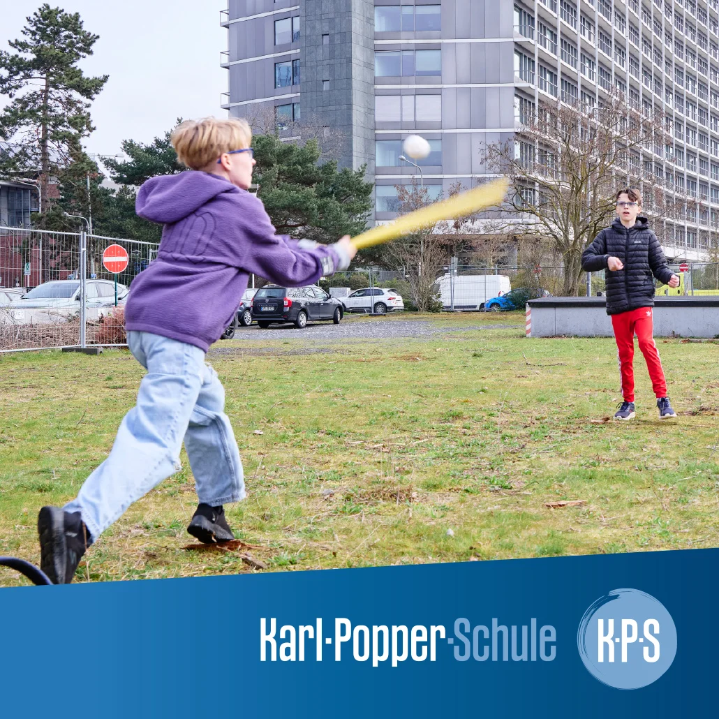 Zwei Kinder spielen auf einer Wiese vor einem Schulgebäude: ein Kind schwingt einen gelben Schläger und trifft einen Ball, das andere steht wartend.