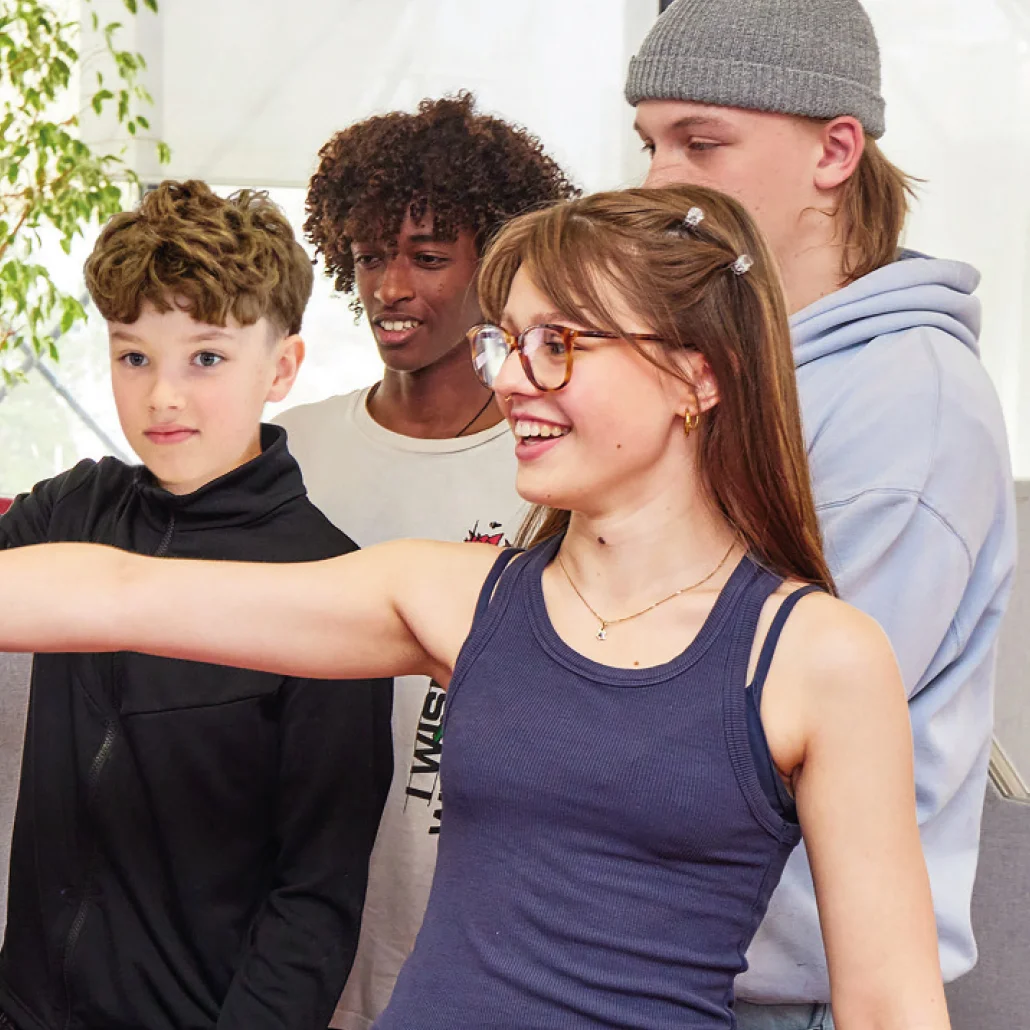 Vier Jugendliche stehen nah beieinander; vorne ein lächelndes Mädchen mit Brille und ausgestrecktem Arm, dahinter drei Jungen, ebenfalls aufmerksam und freundlich.