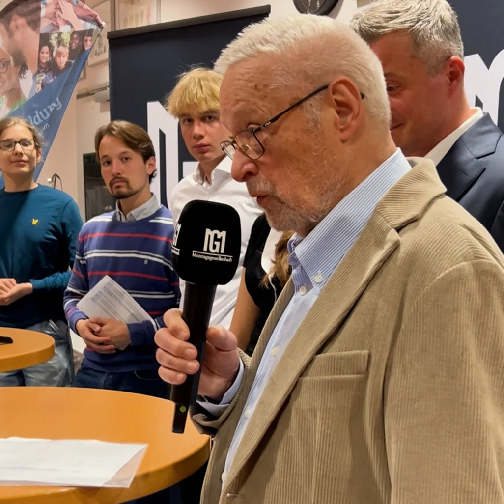 Älterer Mann mit Brille und Mikrofon spricht vor einer kleinen Gruppe junger Erwachsener, die aufmerksam zuhören, in einem Innenraum mit Stehtischen.