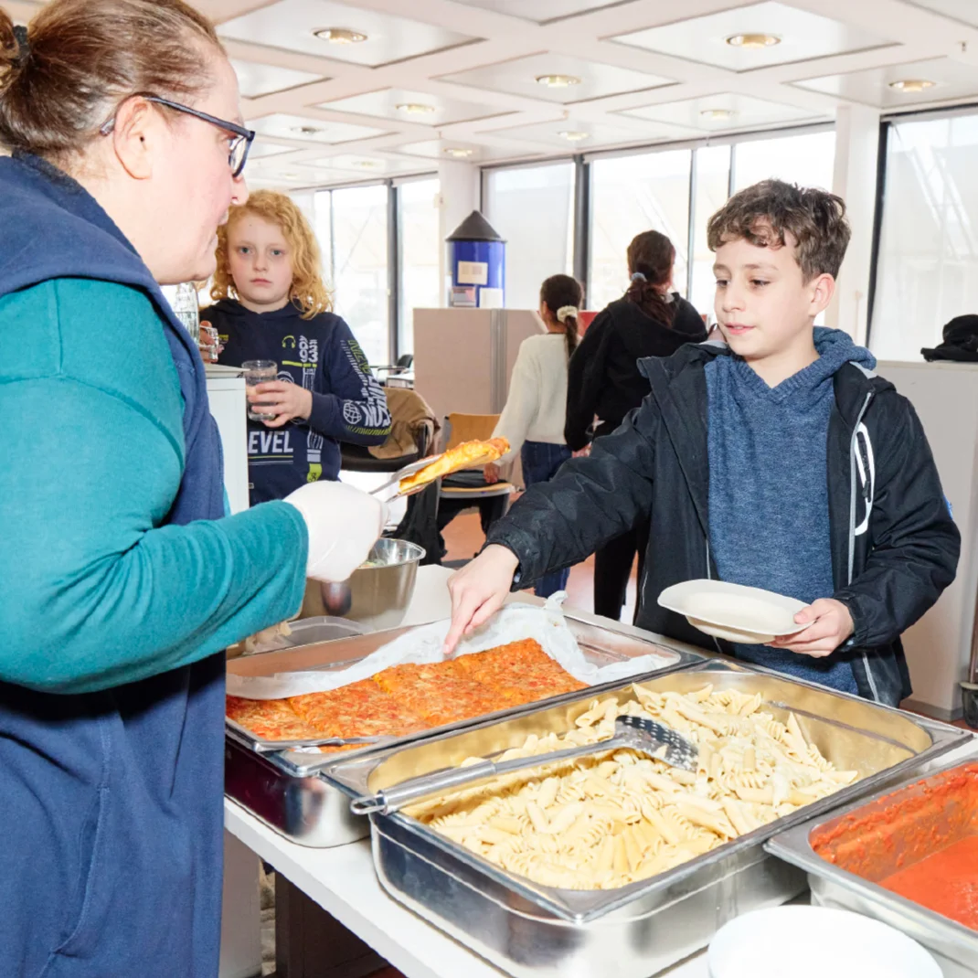 Frau serviert einem Jungen an einer Essensausgabe ein Stück Auflauf; Tabletts mit Pasta und Tomatensauce stehen bereit, weitere Kinder warten in der Schlange.