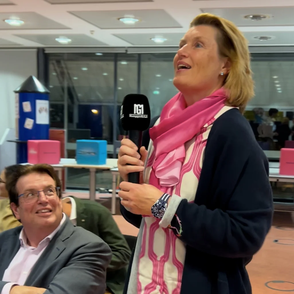 Frau steht in einem Innenraum, spricht in ein Mikrofon und blickt nach oben; sie trägt einen pinken Schal und gemustertes Oberteil, ein Mann sitzt neben ihr und schaut lächelnd zu ihr; Tische und farbige Boxen im Hintergrund.