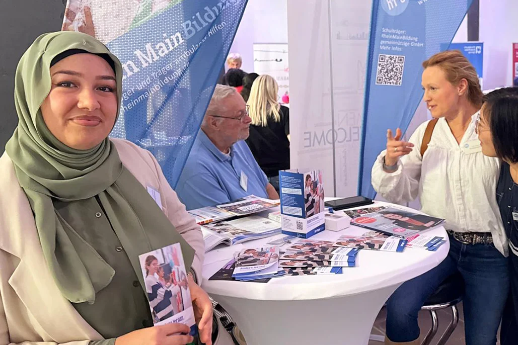 Diverse Messestand-Szene: Junge Frau mit Kopftuch lächelt und hält einen Flyer vor einem Stehtisch mit Broschüren; zwei Frauen unterhalten sich mit einem älteren Mann, Informationsbanner im Hintergrund.