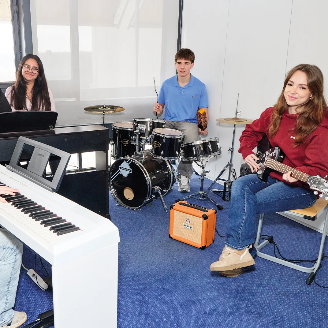 Gruppe junger Musiker in einem Proberaum mit zwei Keyboards, Schlagzeug, E‑Gitarre und kleinem Verstärker, sie sitzen zusammen und proben.
