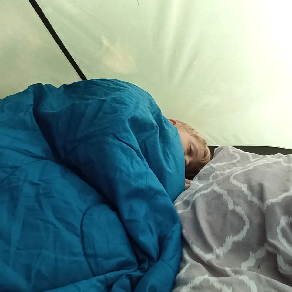 Schlafende Person in blauem Schlafsack liegt in einem Zelt, Kopf auf grauem, gemustertem Kissen